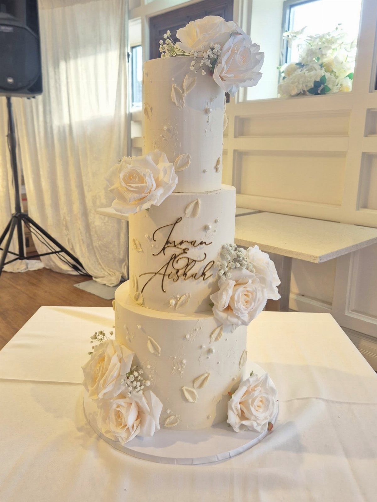 A three-tier white wedding cake adorned with cream roses and gold calligraphy on a white table.