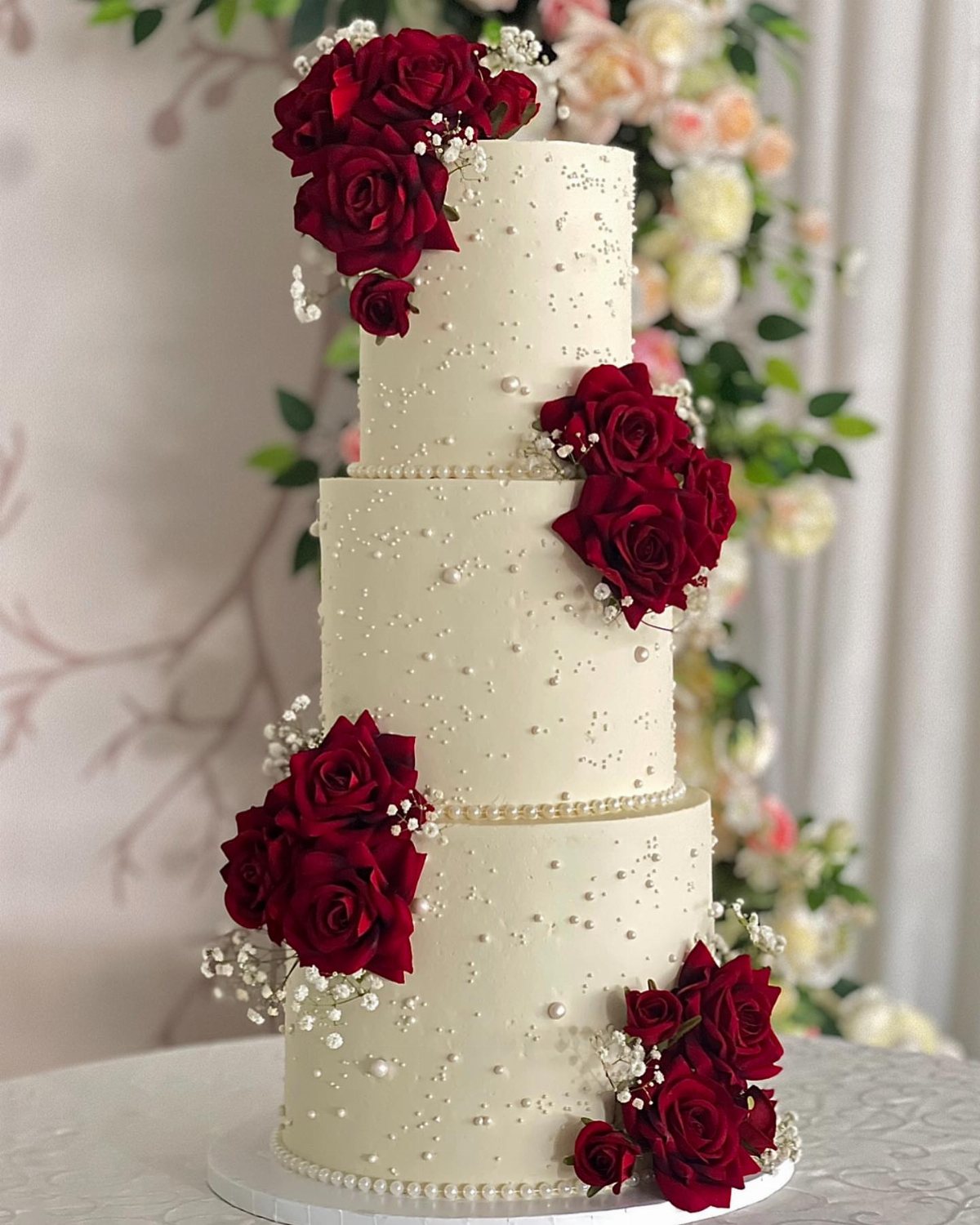 A three-tier white wedding cake adorned with red roses and small white pearls.