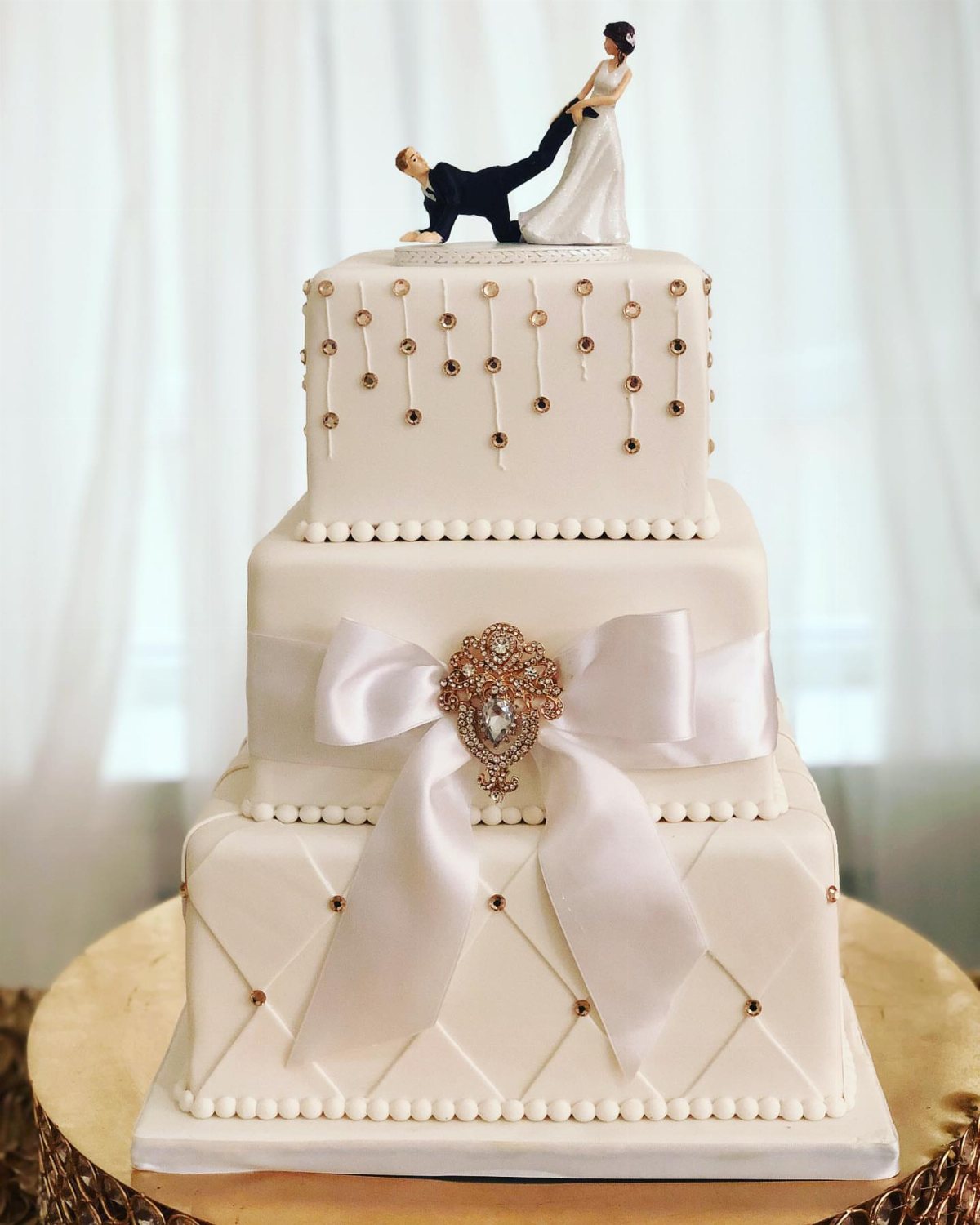 Three-tier square white wedding cake decorated with a large satin bow and gold bead accents.