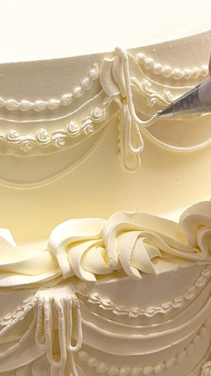 A hand uses a piping tip to decorate a cream wedding cake with white buttercream swirls.