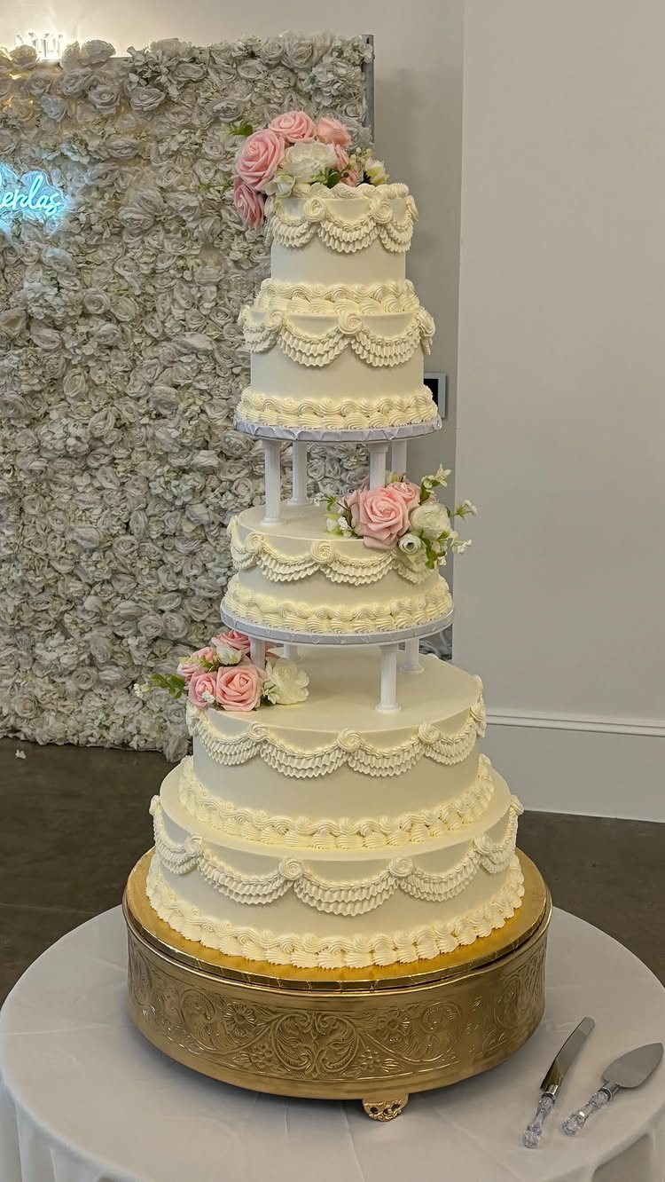 A large white tiered wedding cake with ornate buttercream piping and pink roses on a gold stand.