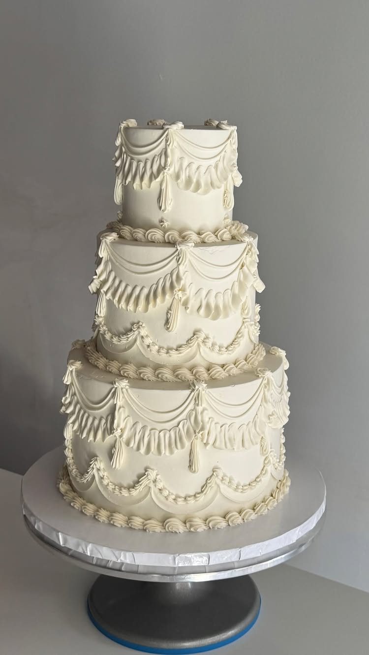 A three-tiered white wedding cake featuring elaborate buttercream piping with scalloped swags and tassels.