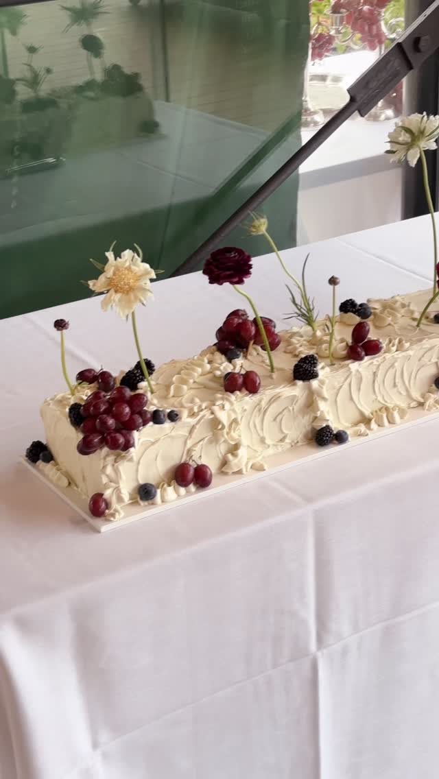 Rectangular white frosted cake with buttercream piping, decorated with grapes, blackberries, and flowers on a white tablecloth.