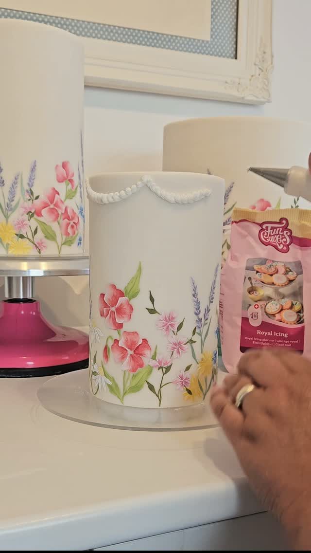 A hand decorating a white floral cake with white royal icing pearls using a piping bag.