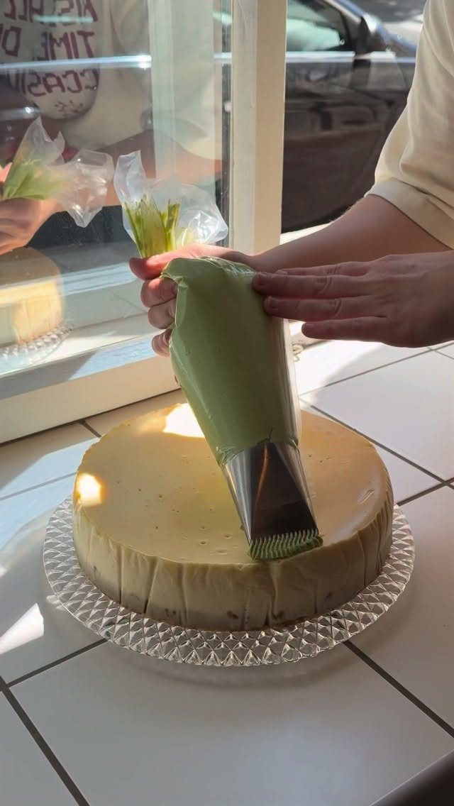 Hands using a green frosting piping bag with a metal tip to decorate a round yellow cake.