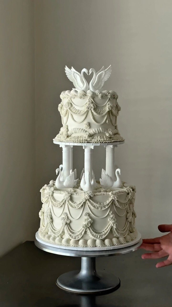 Two-tiered white wedding cake with ornate buttercream piping and swan toppers on a silver stand.
