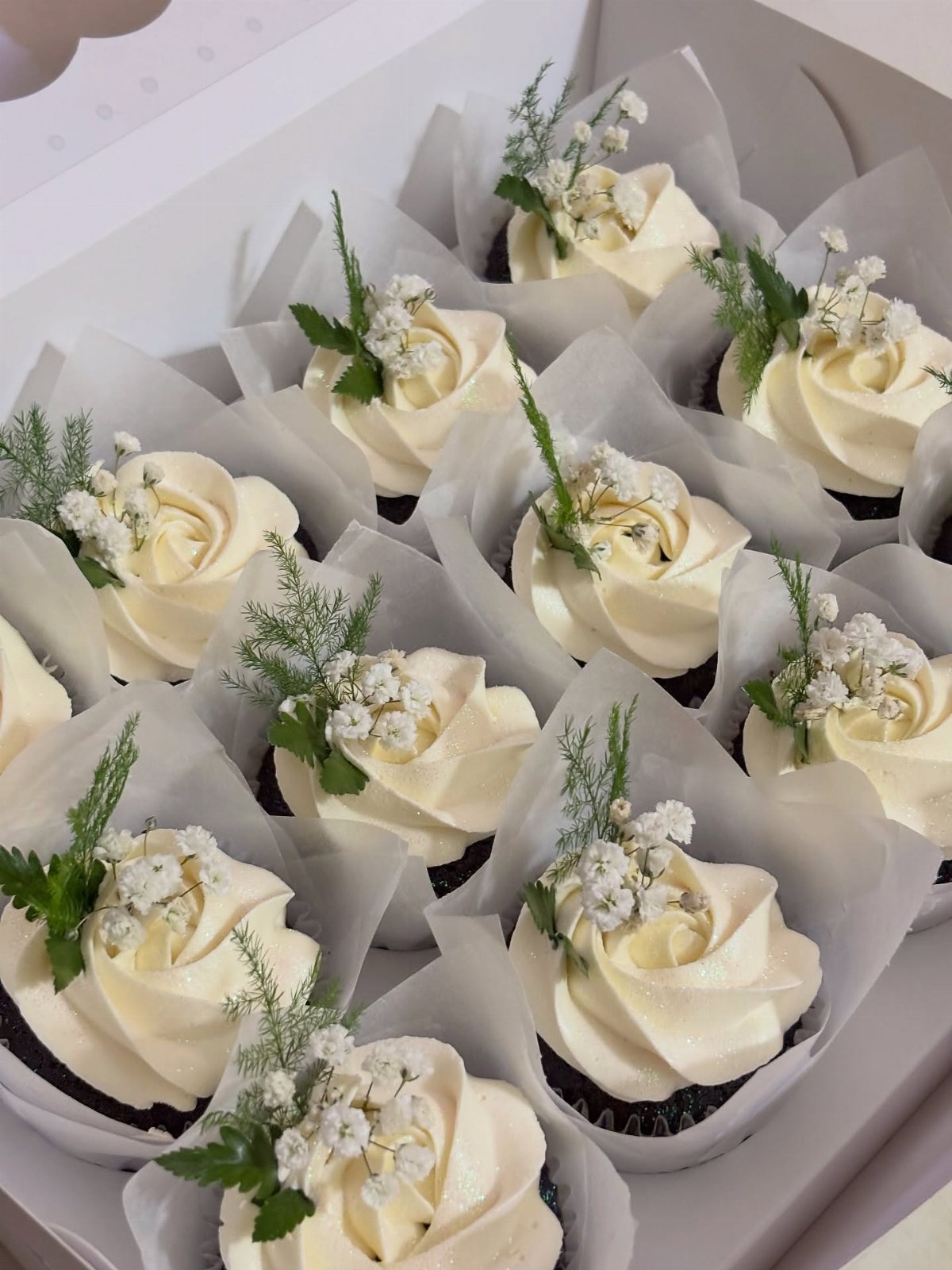 Chocolate cupcakes with white buttercream frosting and small white flowers in a white box.