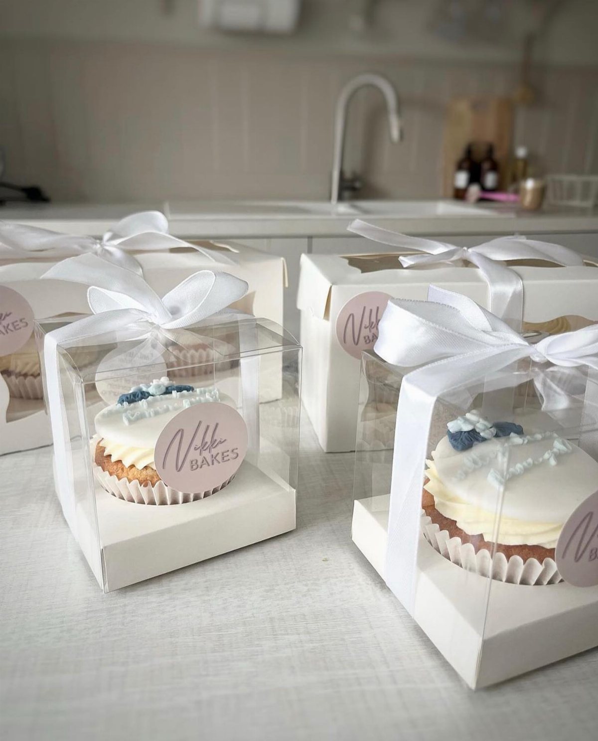 Individual cupcakes with white frosting and blue accents packaged in clear boxes with white ribbons.