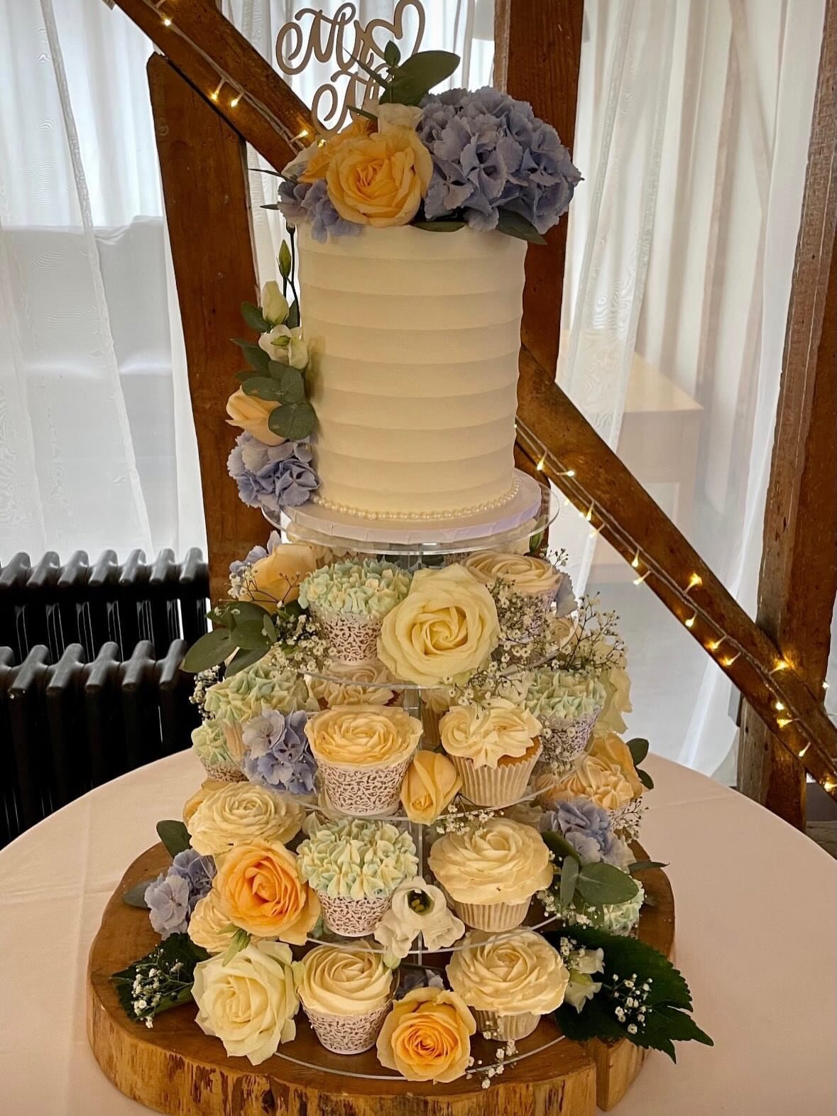 A tiered wedding cake display with a small white cake and various floral cupcakes.