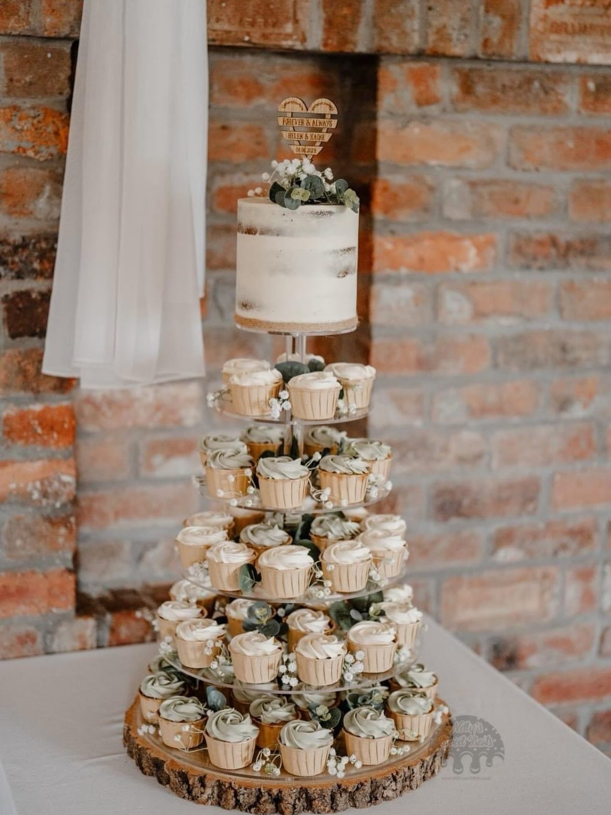 A tiered cupcake tower topped with a small white semi-naked wedding cake on a wooden base.
