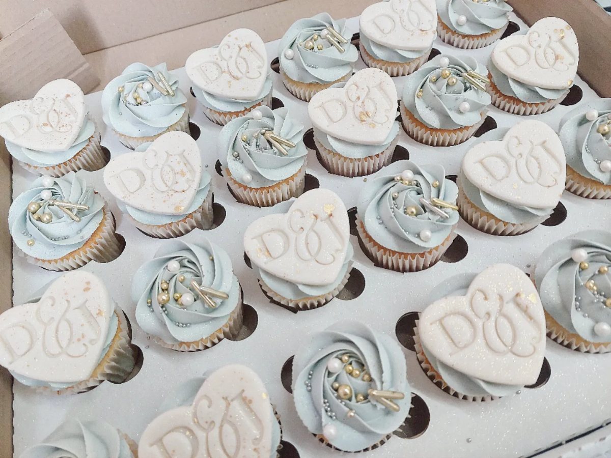 Cupcakes with light blue frosting and white heart-shaped toppers decorated with gold initials and pearls.