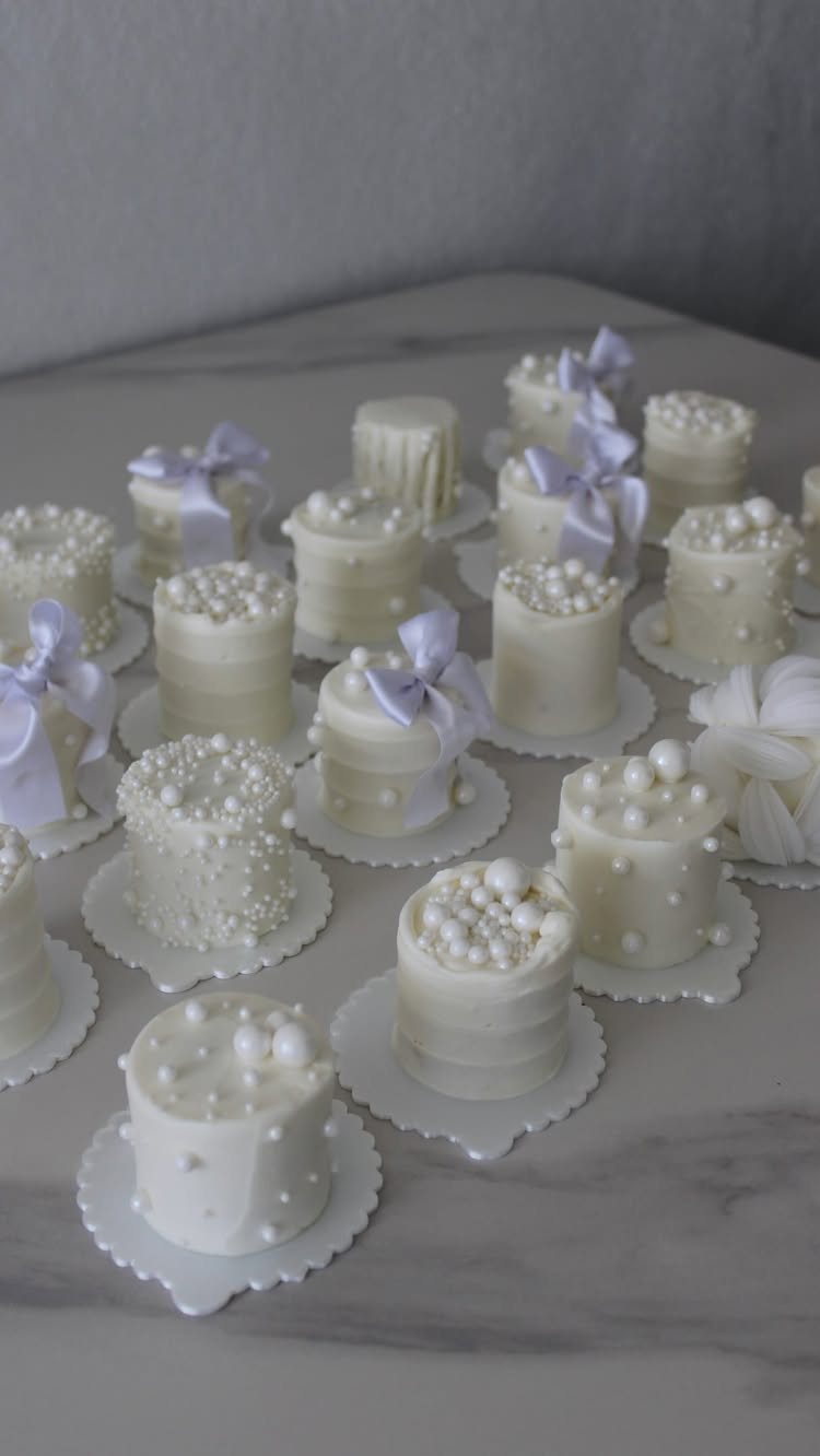 Small white individual cakes decorated with pearls and bows on a marble surface.