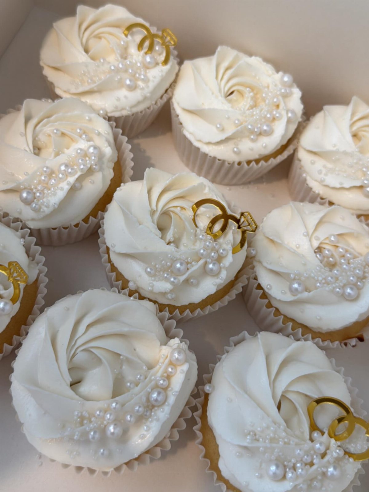 White frosted cupcakes decorated with edible pearls and gold wedding ring toppers in a box.