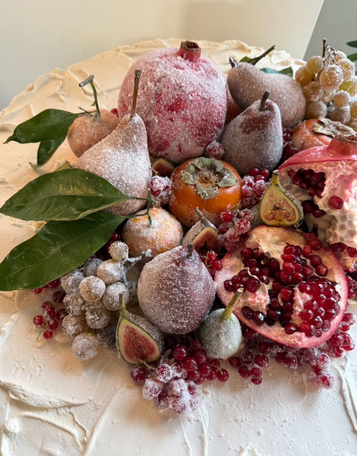 A white frosted cake topped with powdered sugar dusted pears, pomegranates, figs, and grapes.