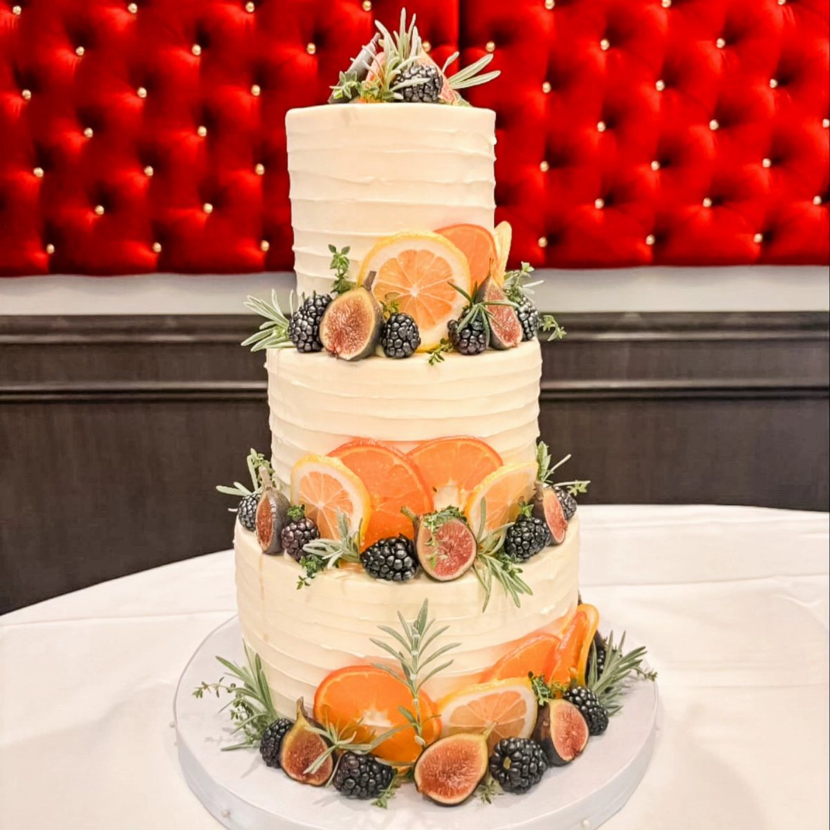 Three-tiered white cake decorated with fresh orange slices, figs, blackberries, and rosemary sprigs.
