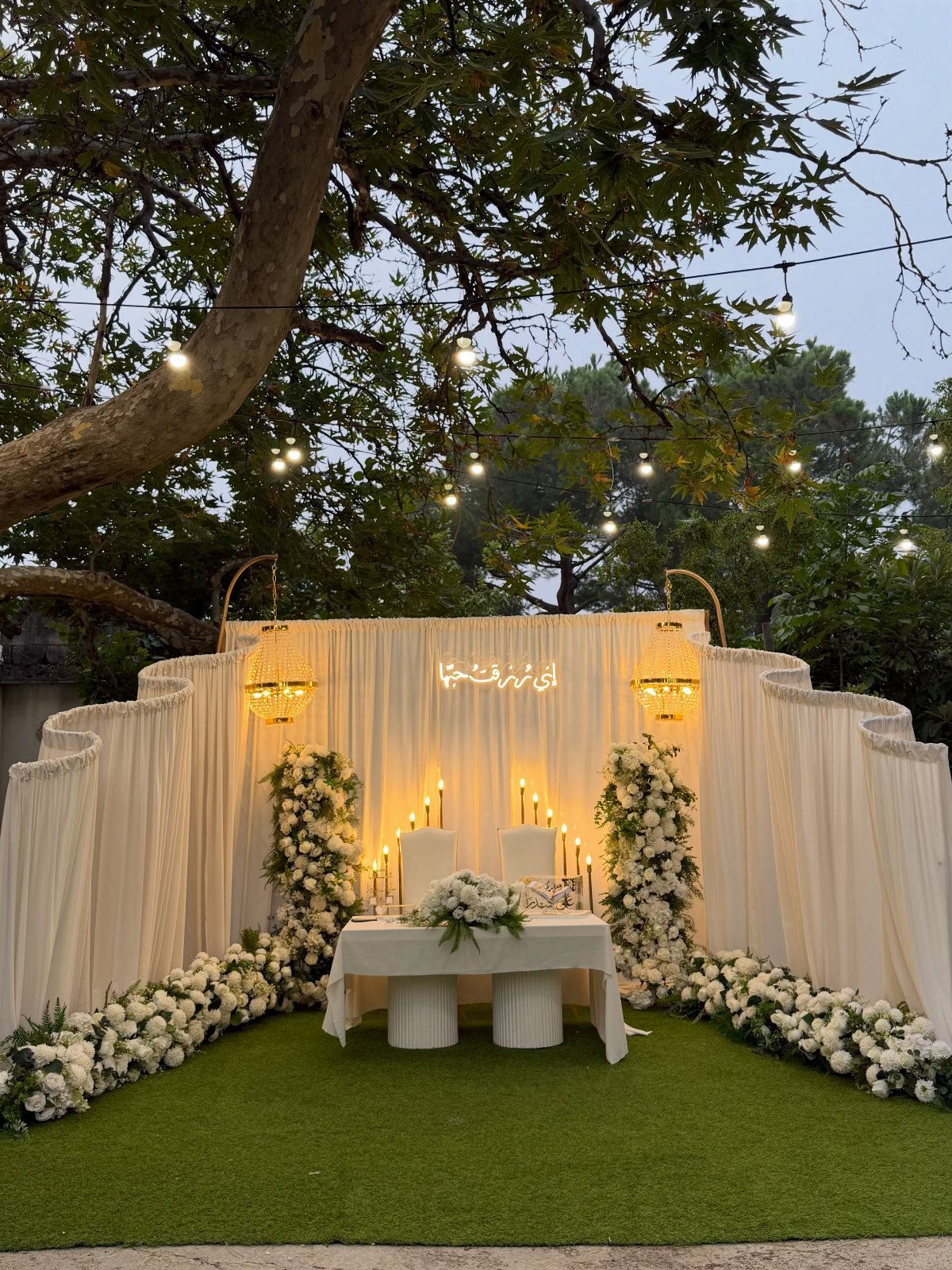 White draped fabric altar with floral arrangements and a central table set on green grass.