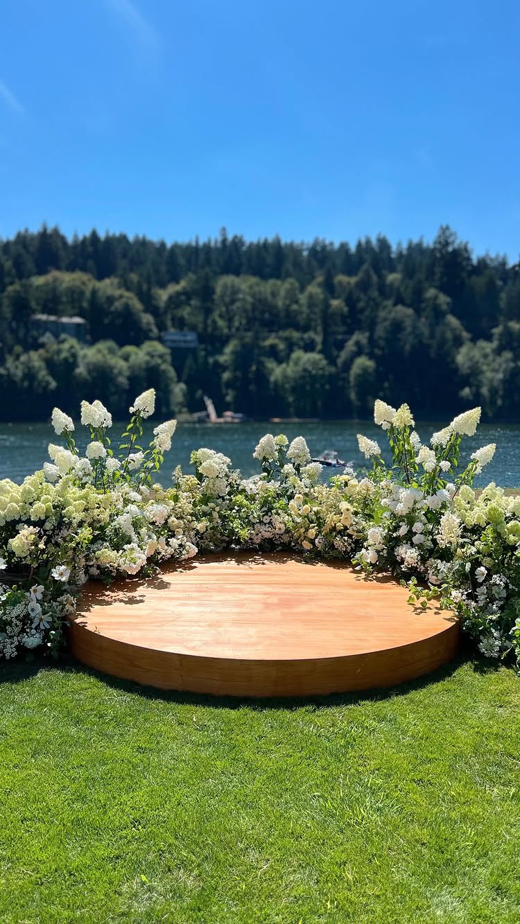 Circular wooden wedding platform surrounded by white floral arrangements on a green lawn by a lake.