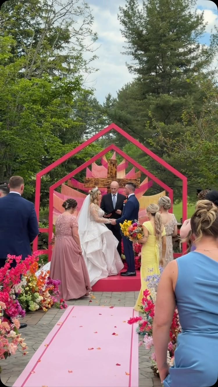 Bride and groom exchanging vows before a large pink geometric wedding altar outdoors.