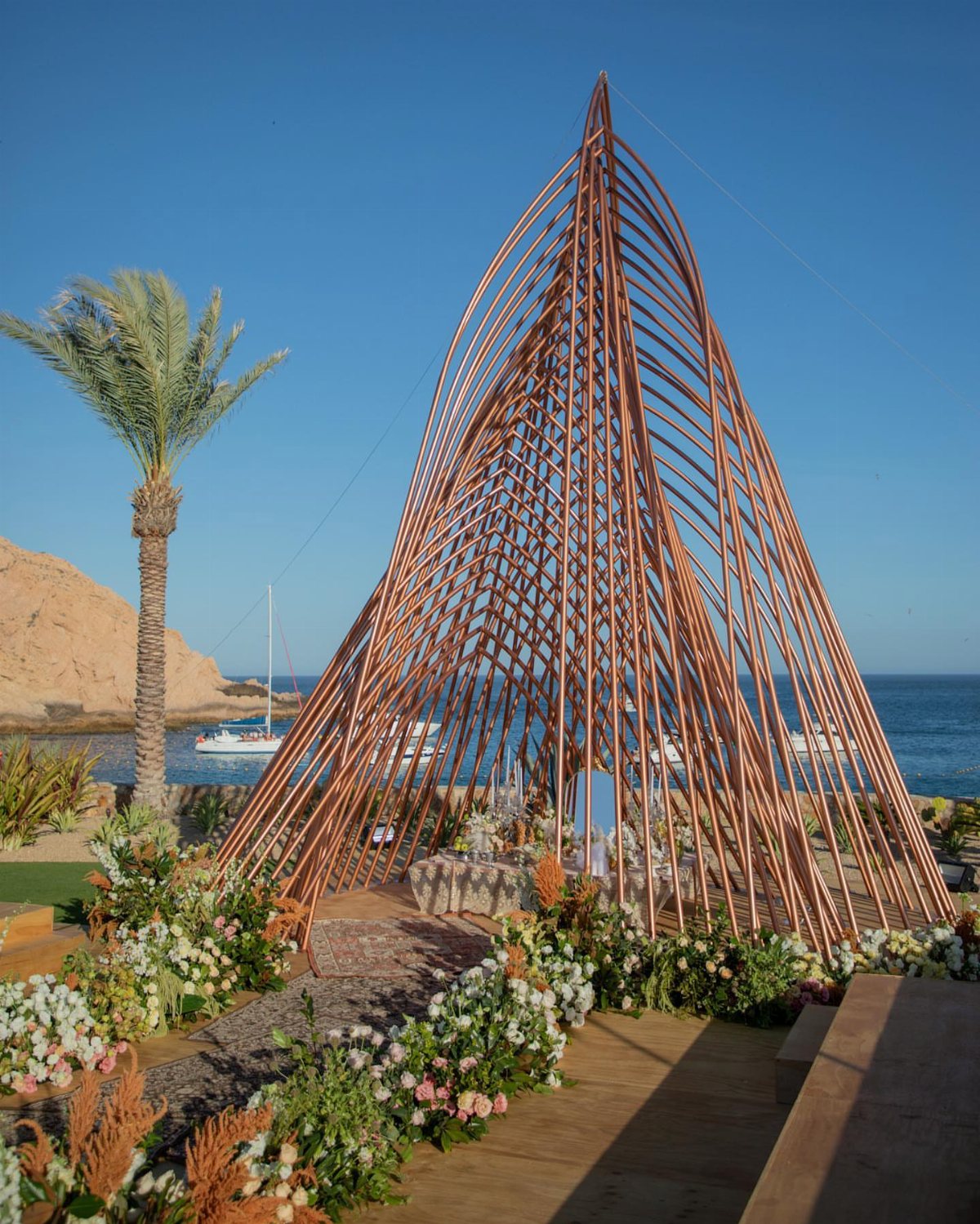 A tall, sculptural copper-colored wedding altar structure overlooking the ocean with white floral arrangements.