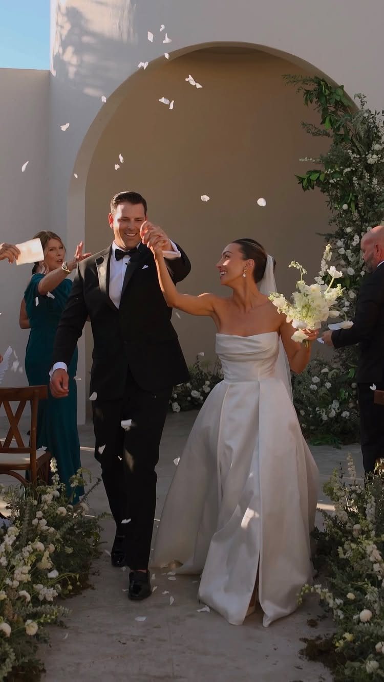 Newlyweds walk joyfully down the aisle through falling white flower petals.