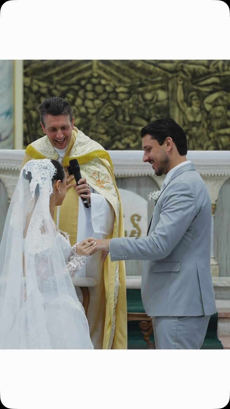 Bride and groom exchanging rings during a wedding ceremony with a priest standing behind them.