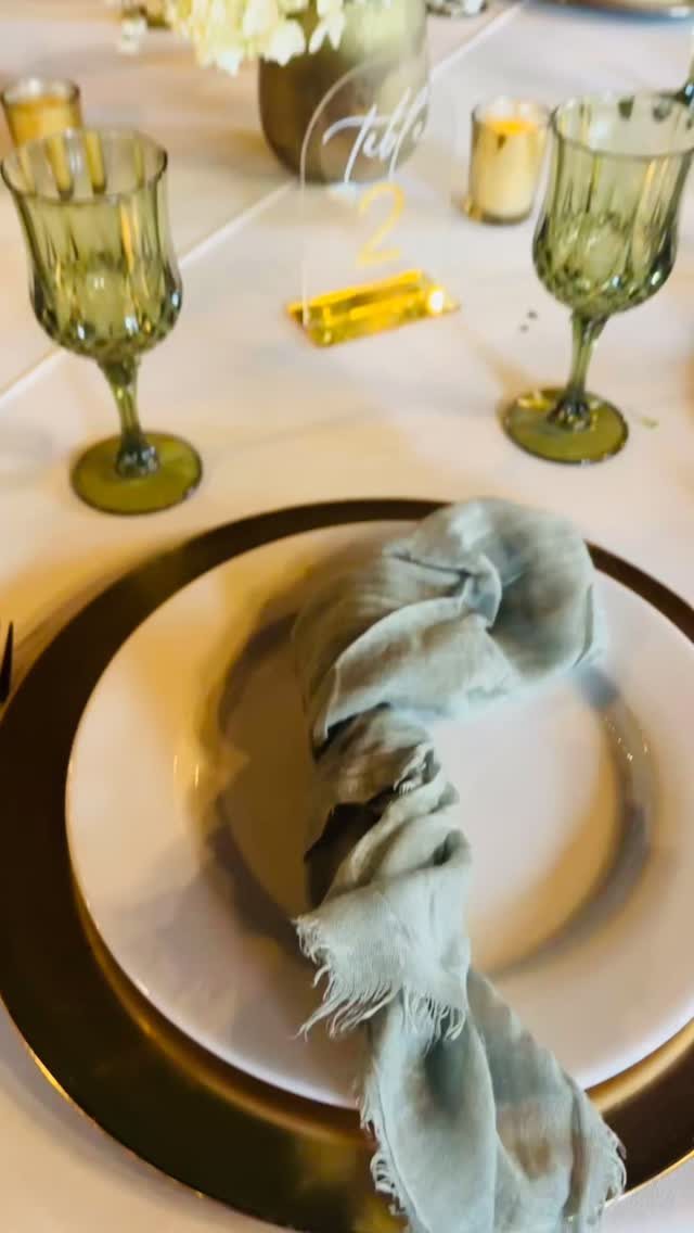 Dusty blue napkin on a white plate with gold charger and green glassware on a table.