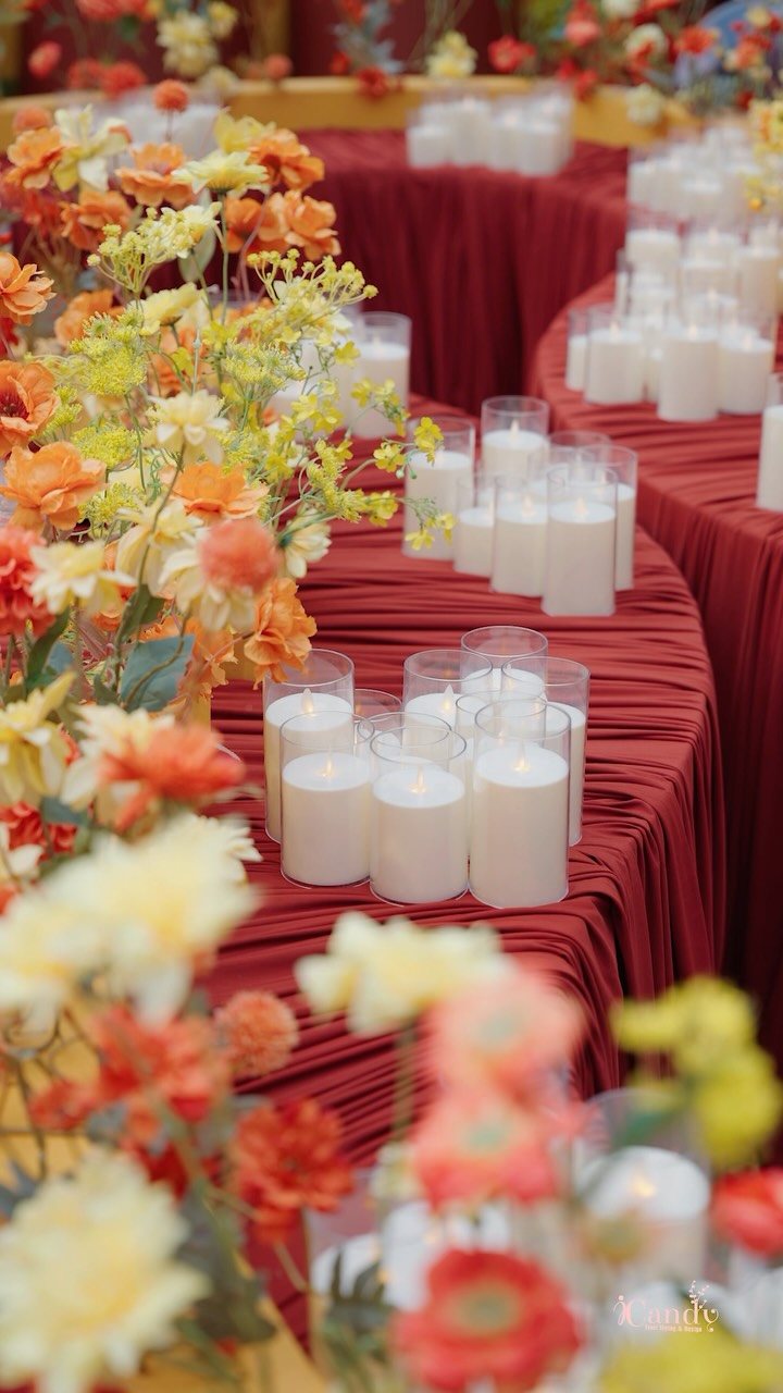 Floral arrangements in orange and yellow tones sit beside white candles on a red tablecloth.