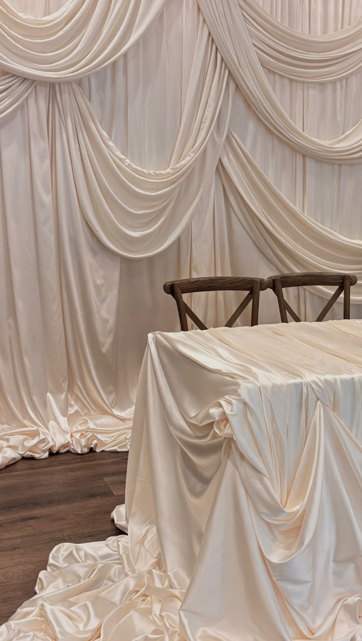 Cream satin fabric draped in elegant swags forming a backdrop behind a cloth-covered table and wooden chairs.