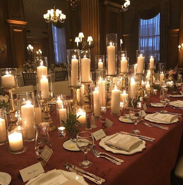 Long dining table decorated with many tall white pillar candles in glass cylinders on a red cloth.