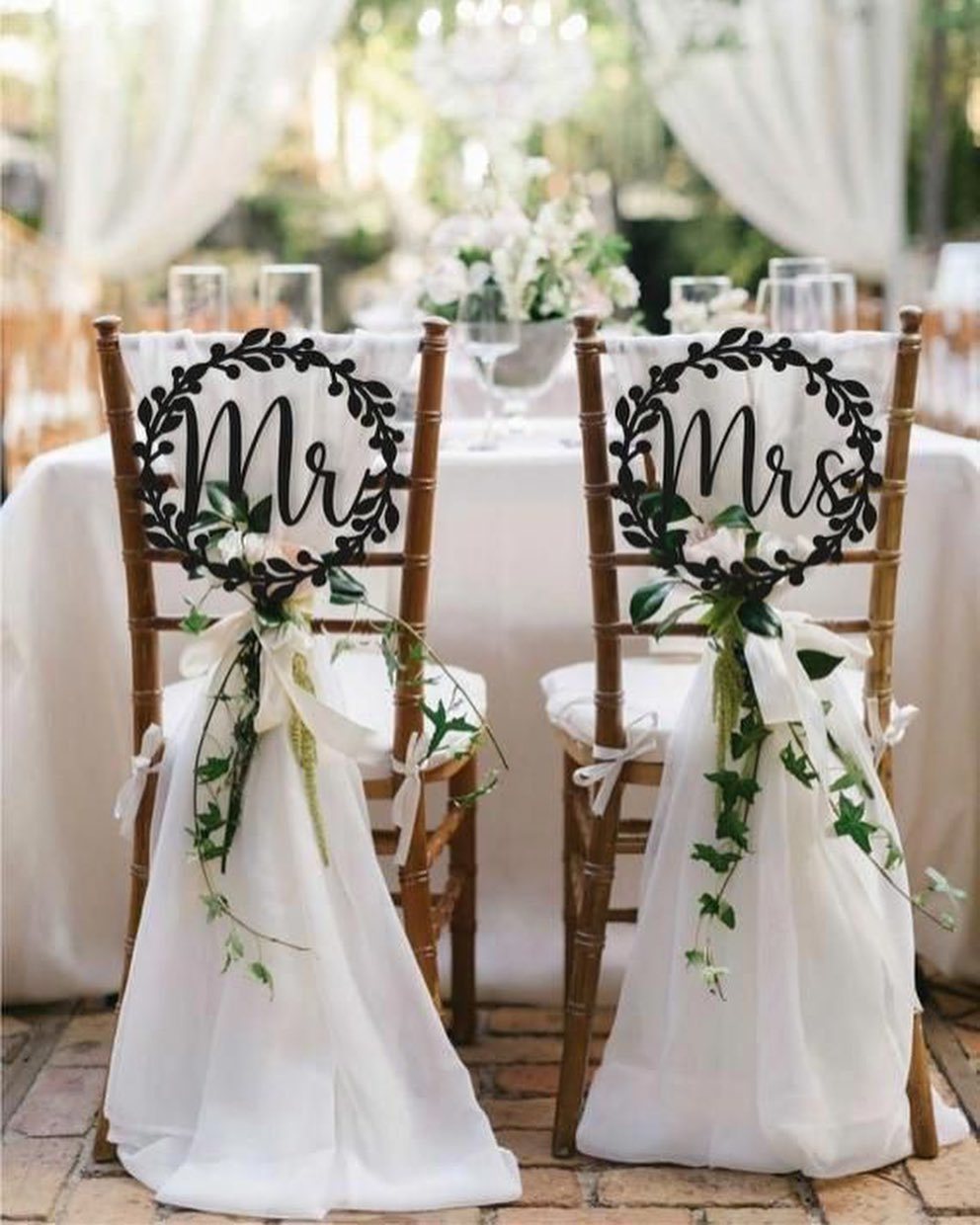 Two wooden wedding chairs decorated with white fabric, greenery, and Mr and Mrs signs.