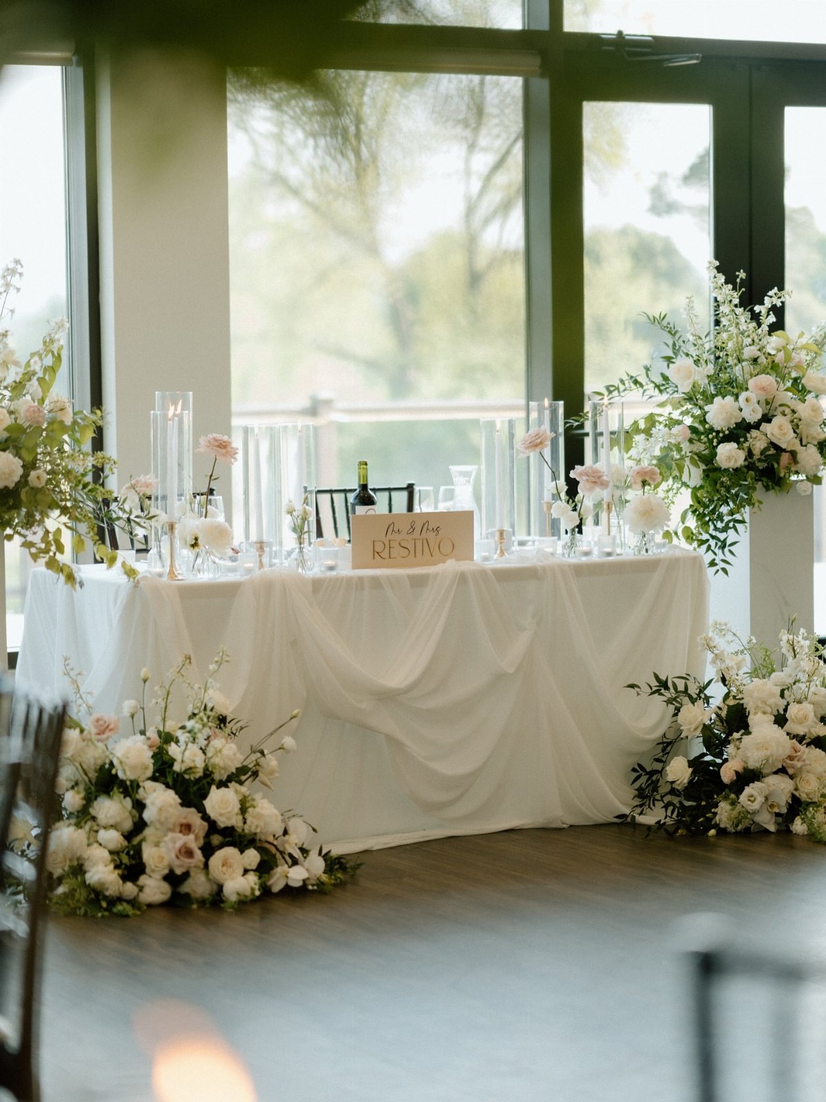 A white draped wedding head table decorated with lush white and pink floral arrangements and tall glass candles.