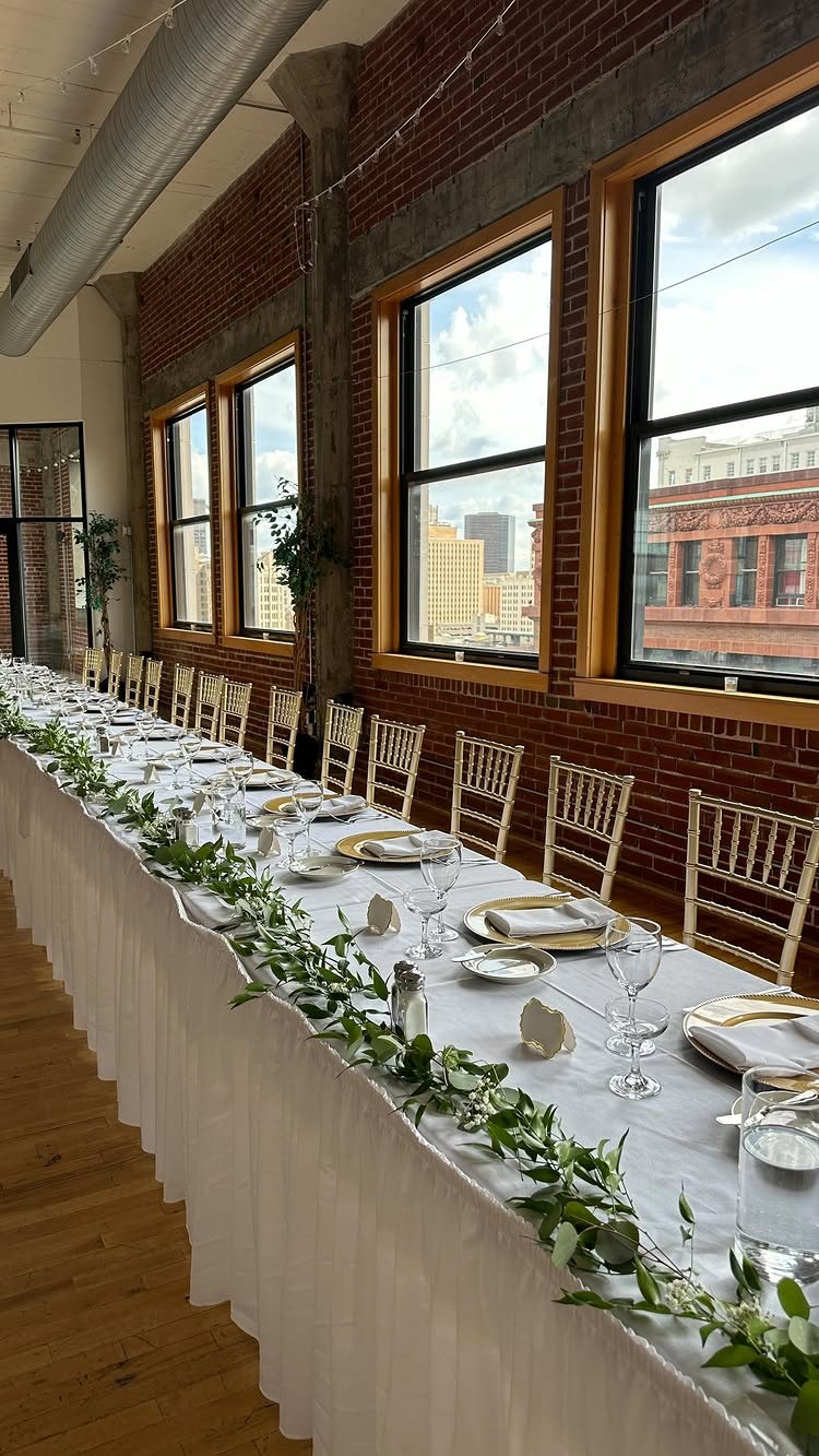 A long wedding head table decorated with white linens, gold plates, and a green leafy garland.