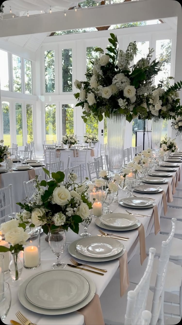 A long wedding dining table decorated with white flowers, candles, and white plates in a bright room.