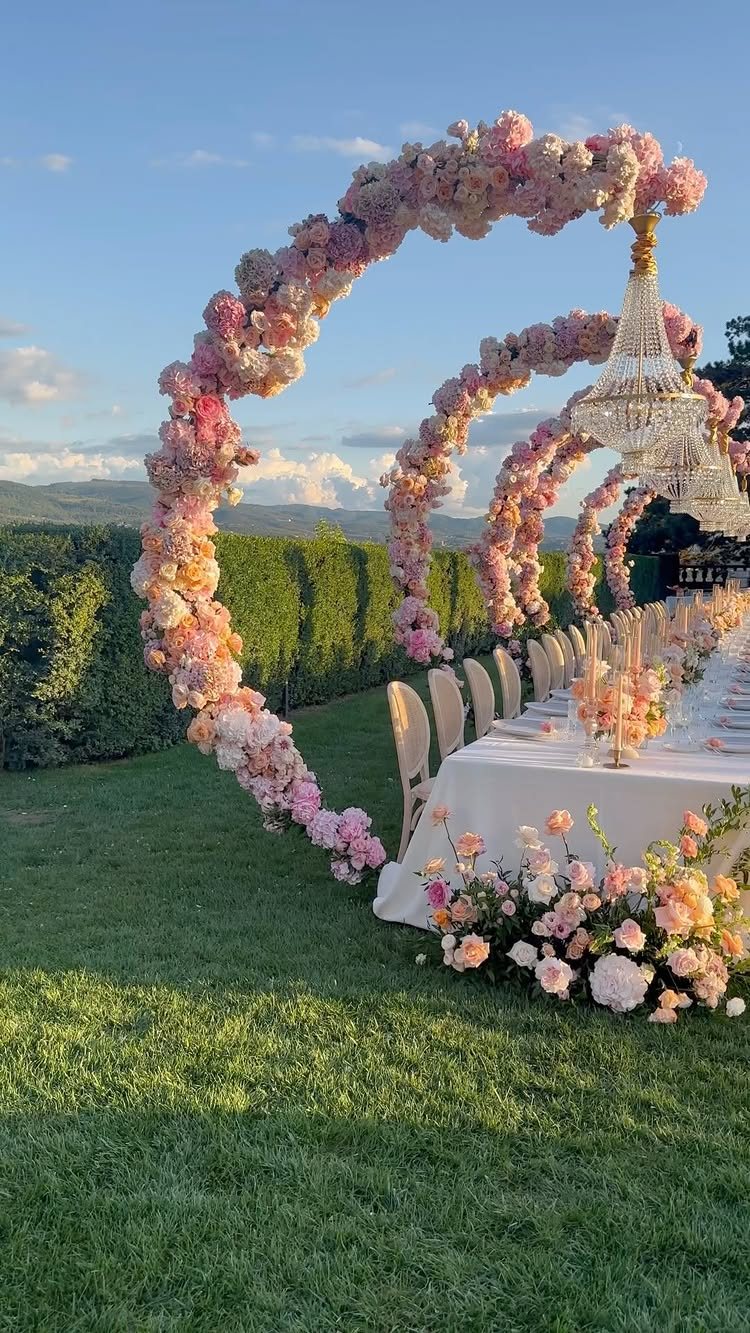 A long wedding head table decorated with pink floral arches and hanging crystal chandeliers outdoors.