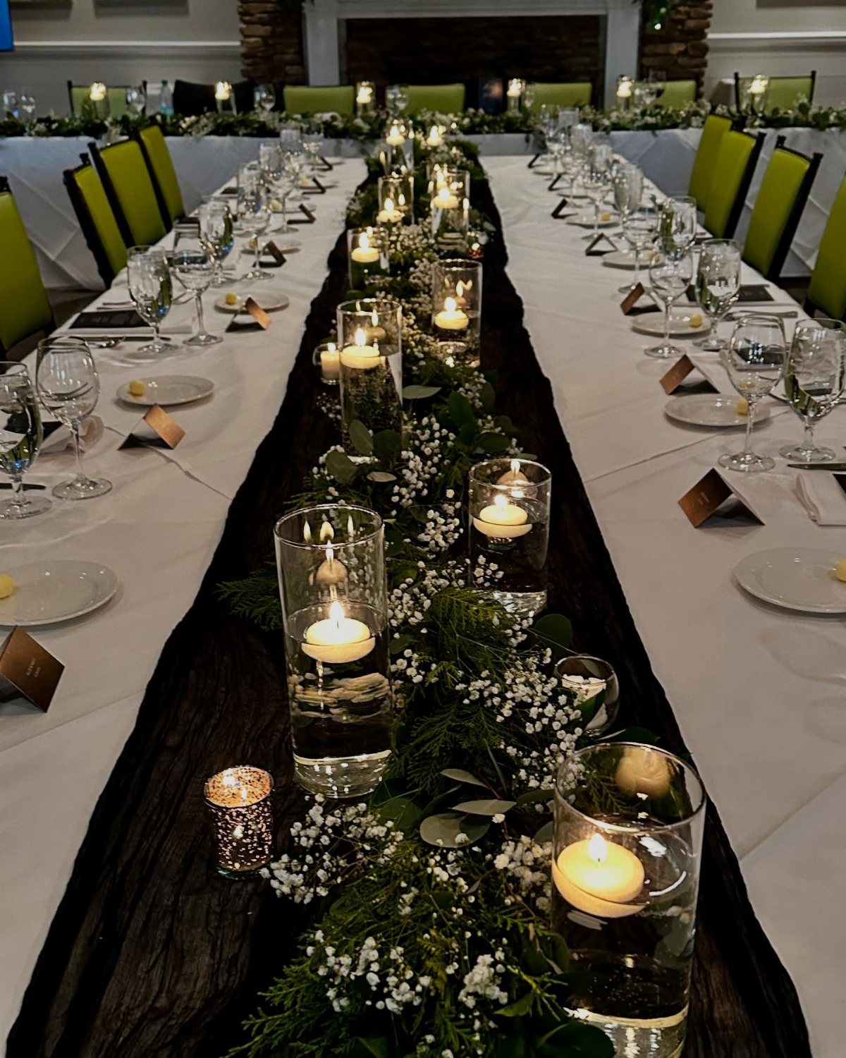 Dark textured table runner decorated with greenery, white flowers, and floating candles on a long wedding table.
