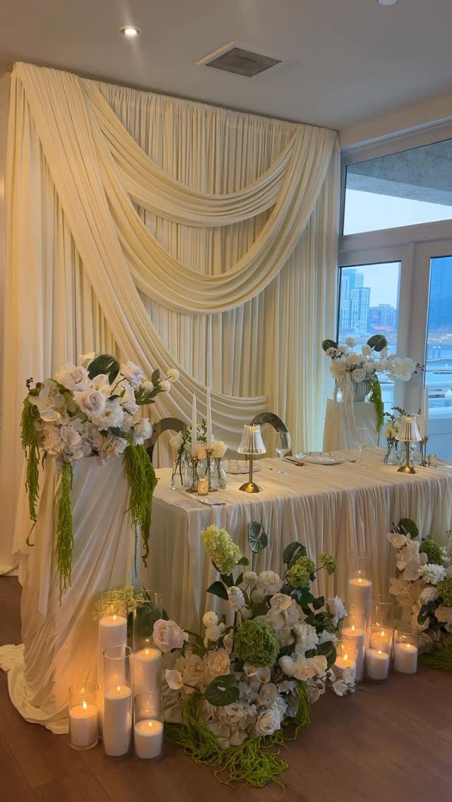 A decorated sweetheart table with white flowers, candles, and elegant cream drapery in the background.