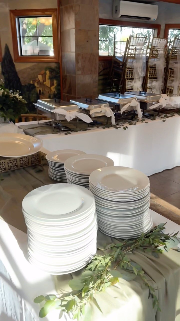 Stacks of white ceramic plates and silver chafing dishes arranged on a long white tablecloth.