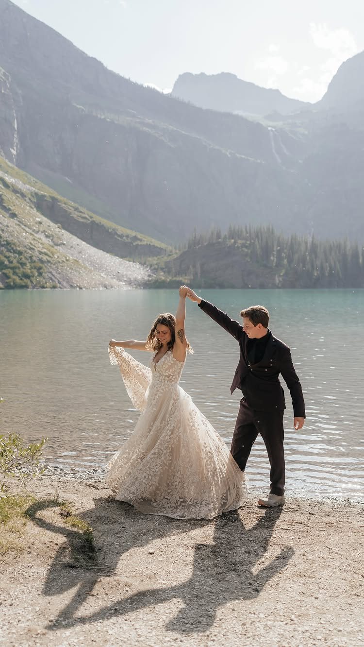 A bride and groom dancing joyfully on a rocky lakeshore with mountains in the background.
