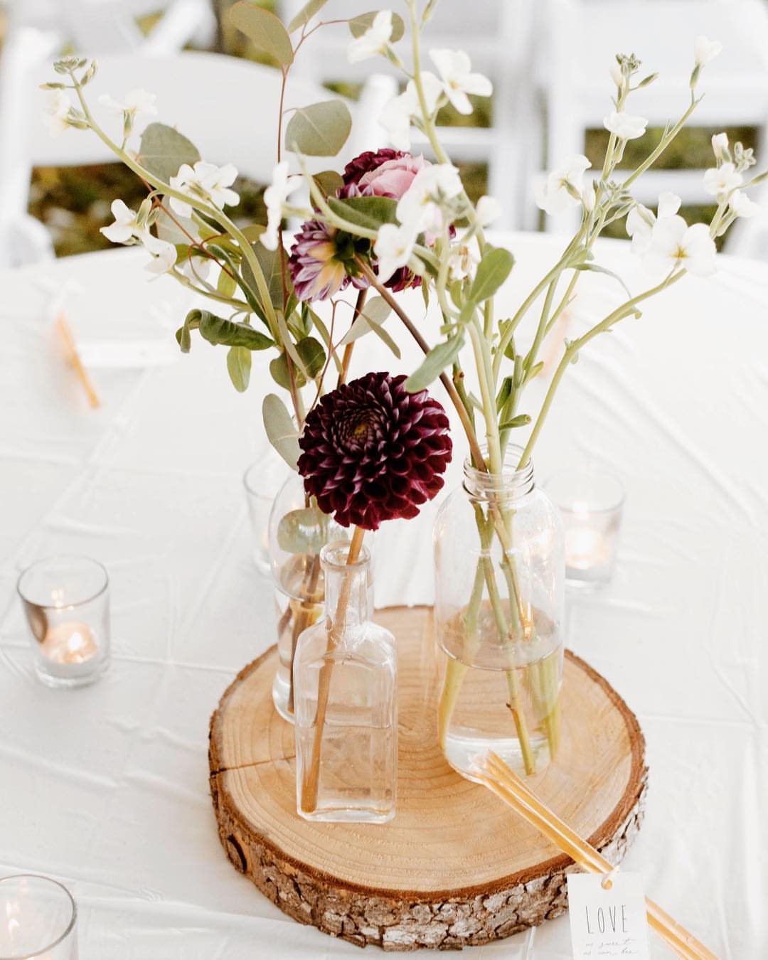 White and burgundy flowers in glass bottles arranged on a wood slice centerpiece.