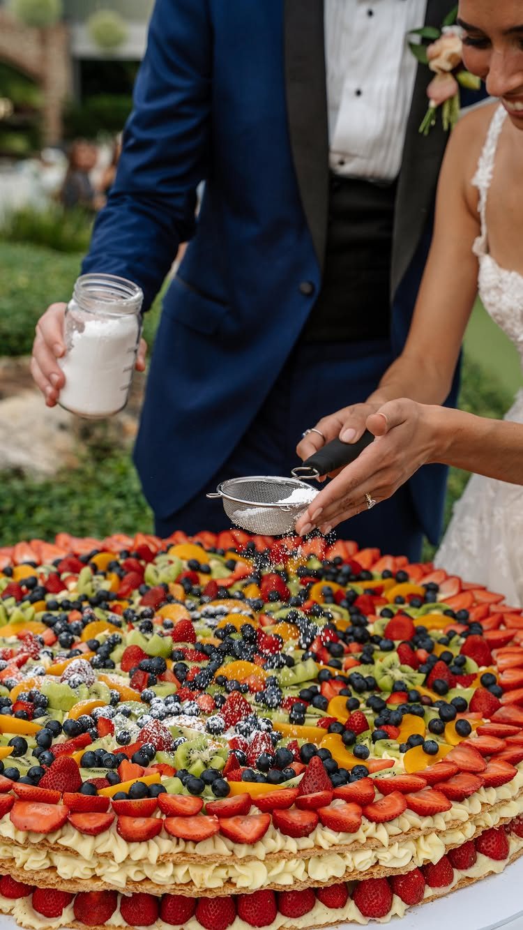 A bride dusting powdered sugar over a large, colorful fruit-topped cake at a wedding.