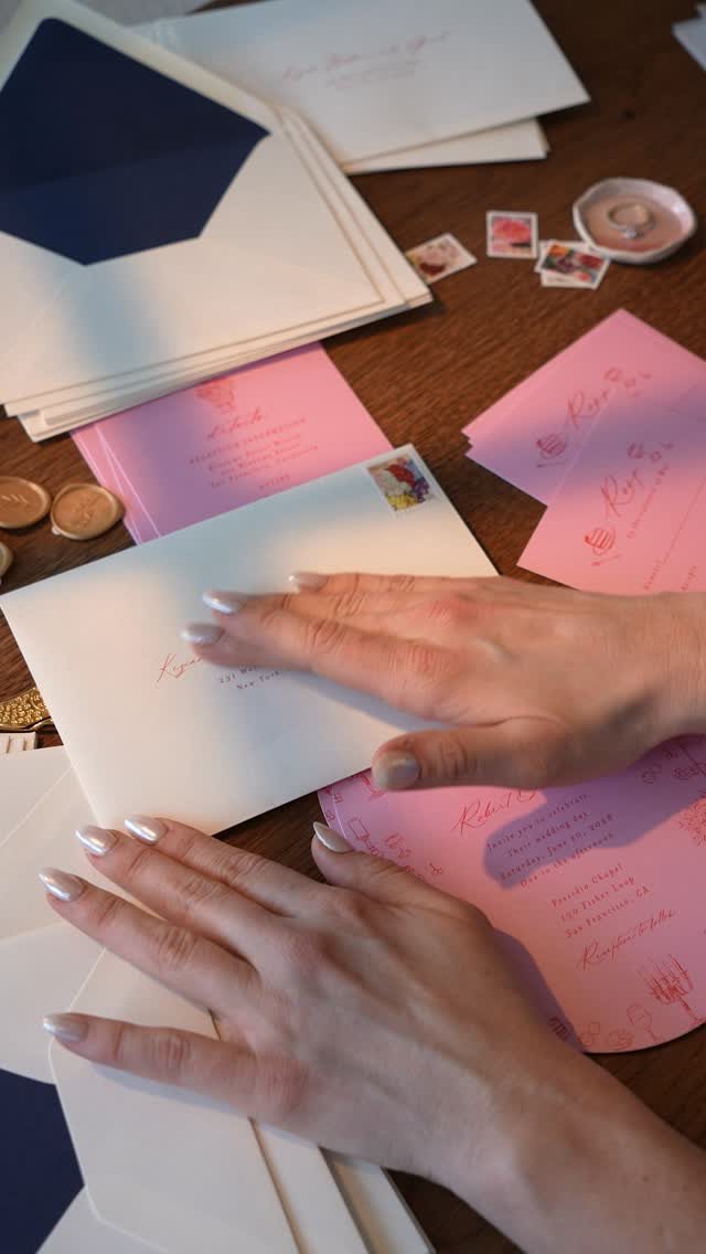 Hands placing stamps on white and pink wedding envelopes on a wooden table.