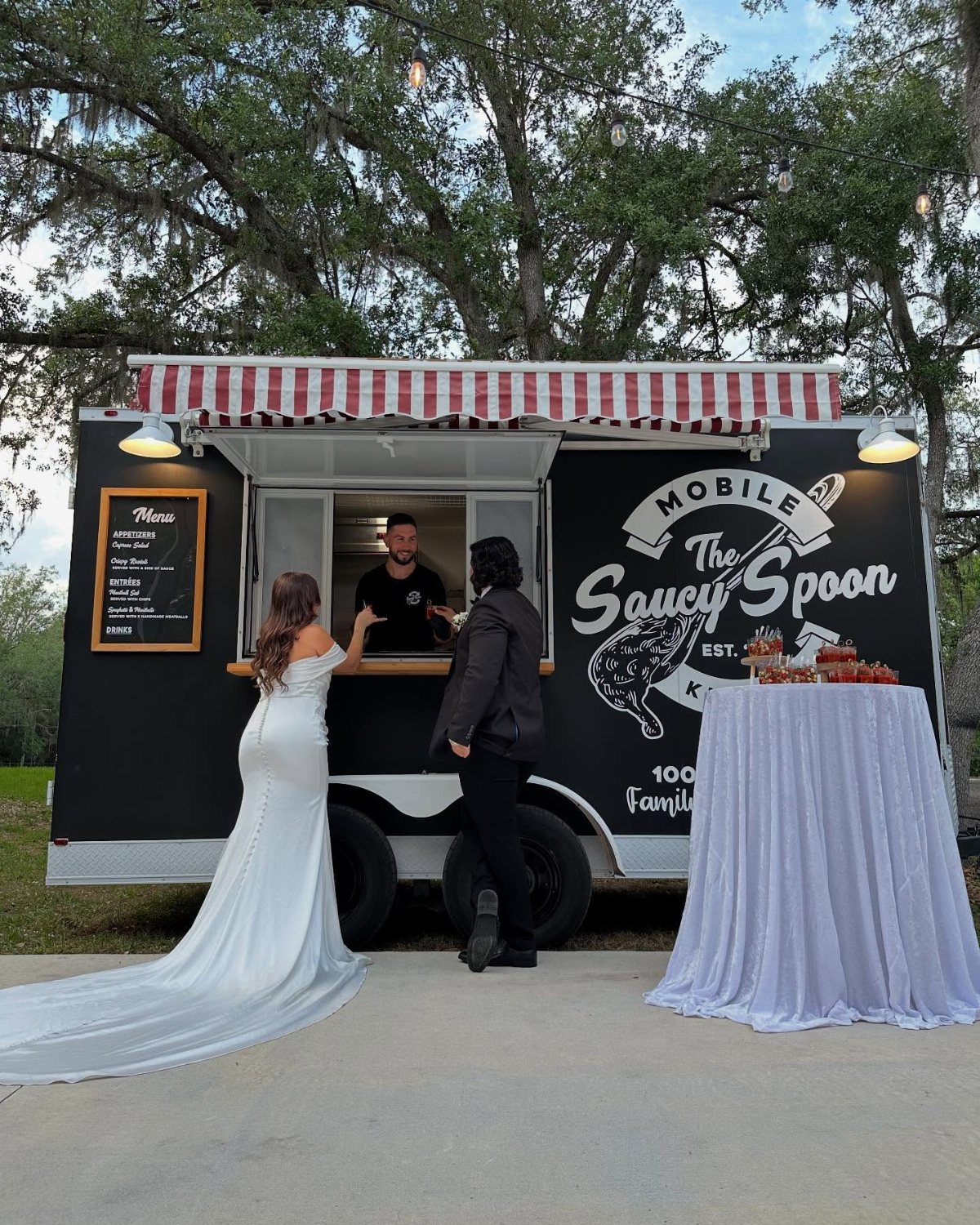 A bride and guest standing before a black mobile food truck under large trees.