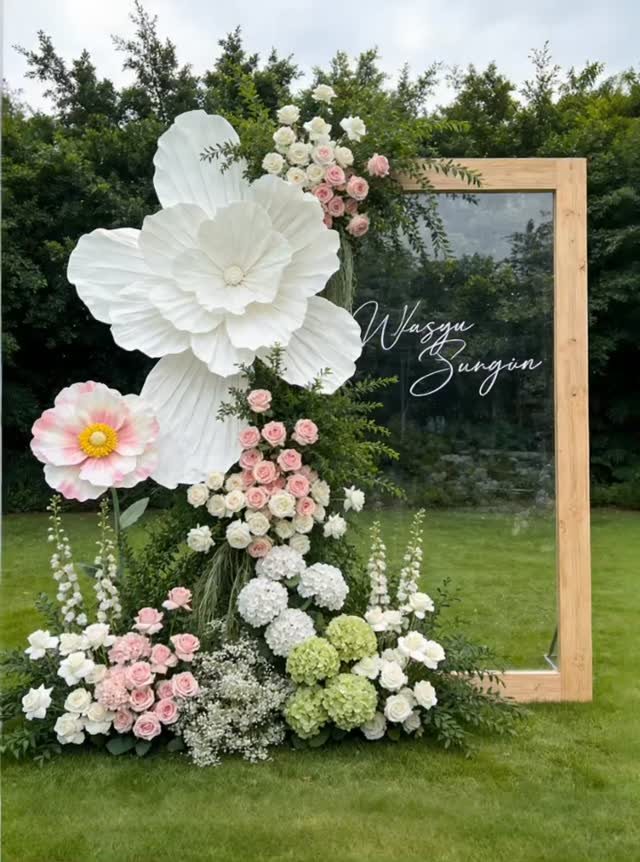 Large white and pink floral arrangement decorating a wooden framed glass wedding sign on a lawn.