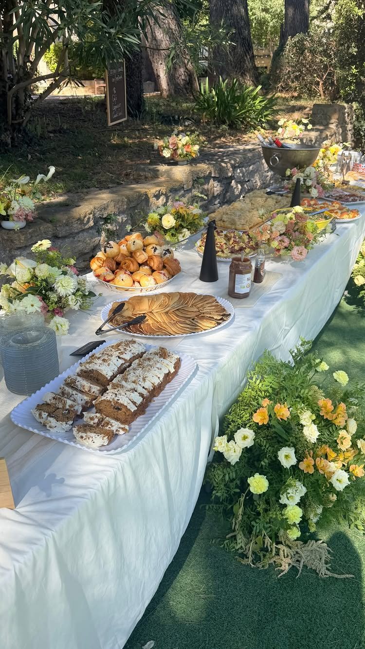 A long buffet table outdoors featuring various pastries, muffins, and cakes decorated with flowers.