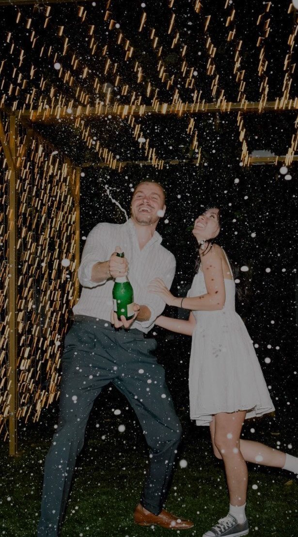 A man spraying champagne from a bottle while a woman in a white dress laughs beside him.