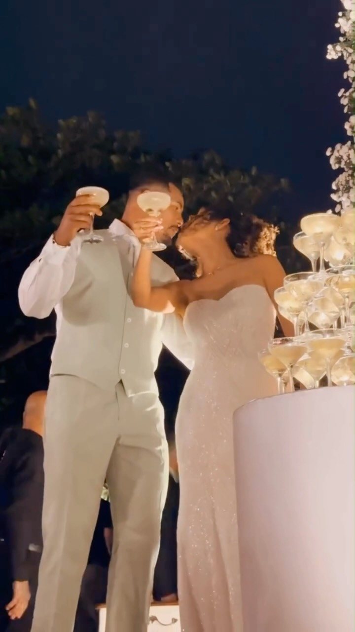 A bride and groom toast with champagne glasses next to a champagne tower at night.