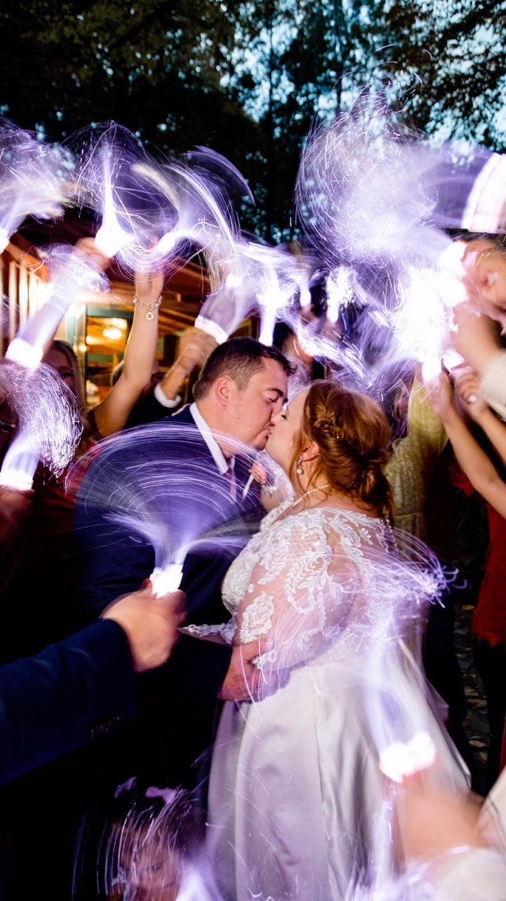 A bride and groom kissing amidst guests waving glowing white fiber optic wands at night.