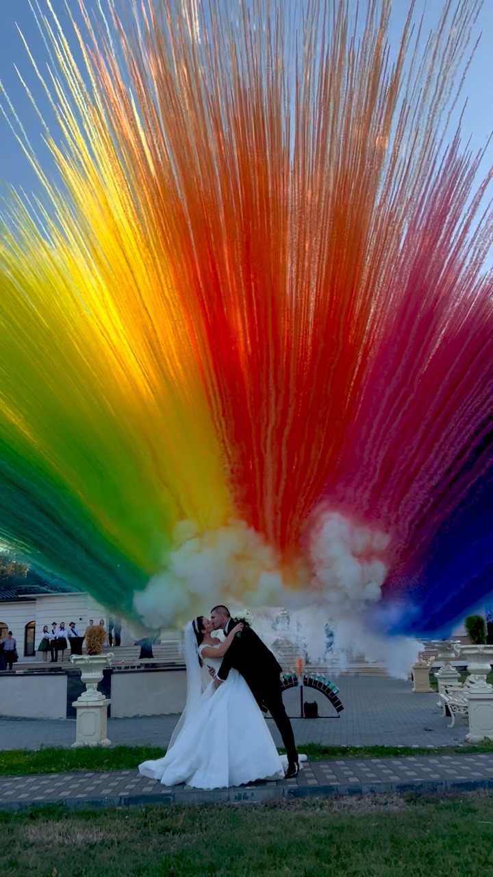 A bride and groom kissing amidst a large, colorful explosion of rainbow-colored smoke.