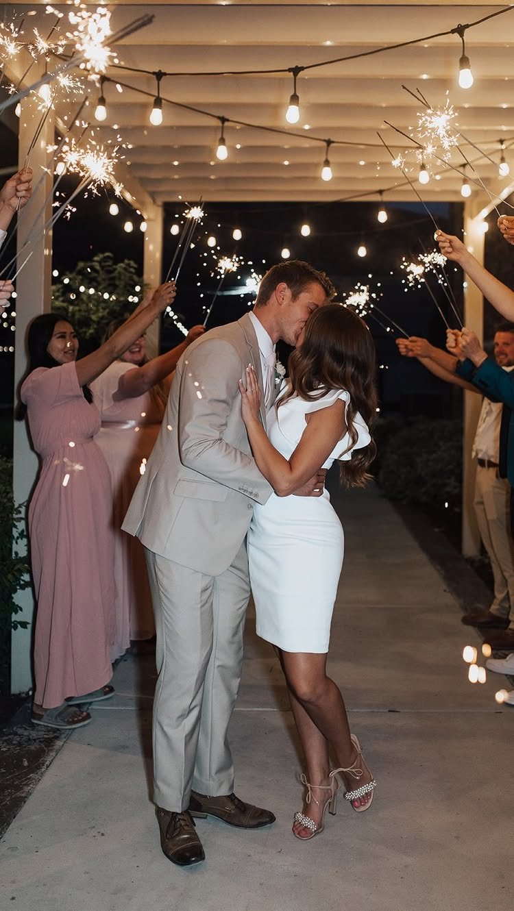 A bride and groom kissing while guests hold glowing sparklers in a nighttime wedding setting.