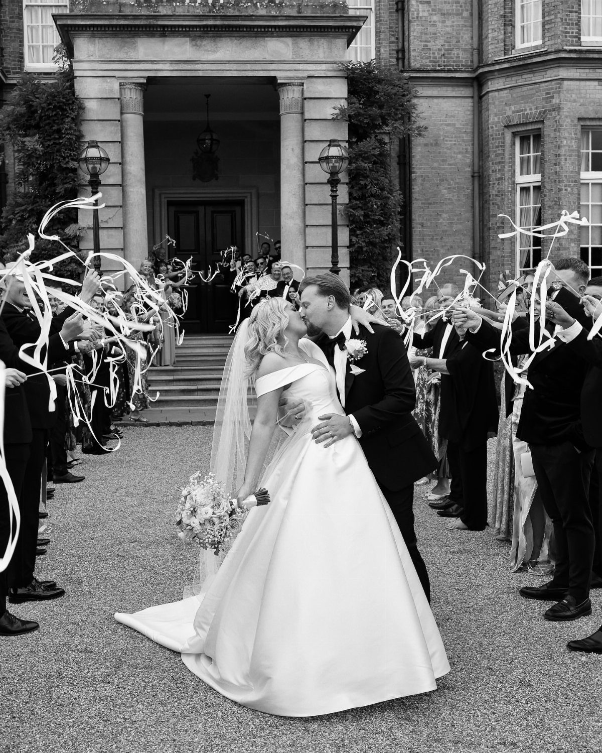 Bride and groom kissing while guests wave white wedding streamers in a courtyard.